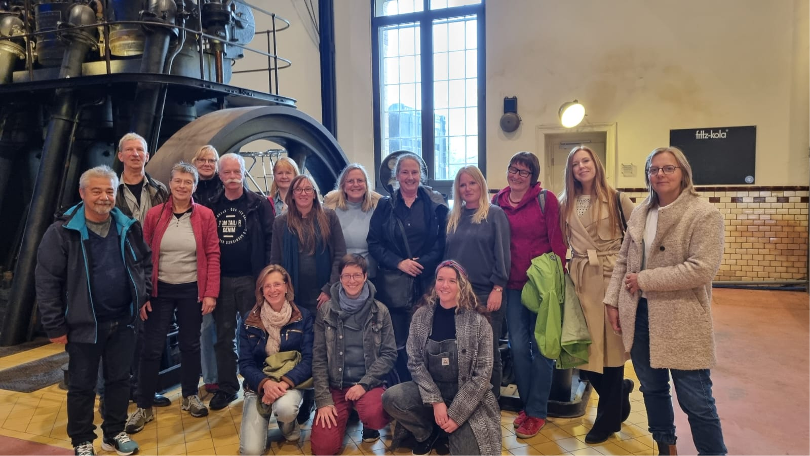 re-use-hessen-14-netzwerktreffen Eine Gruppe von 16 Menschen steht in einer ehemaligen Fabrikhalle vor einer alten Maschine.