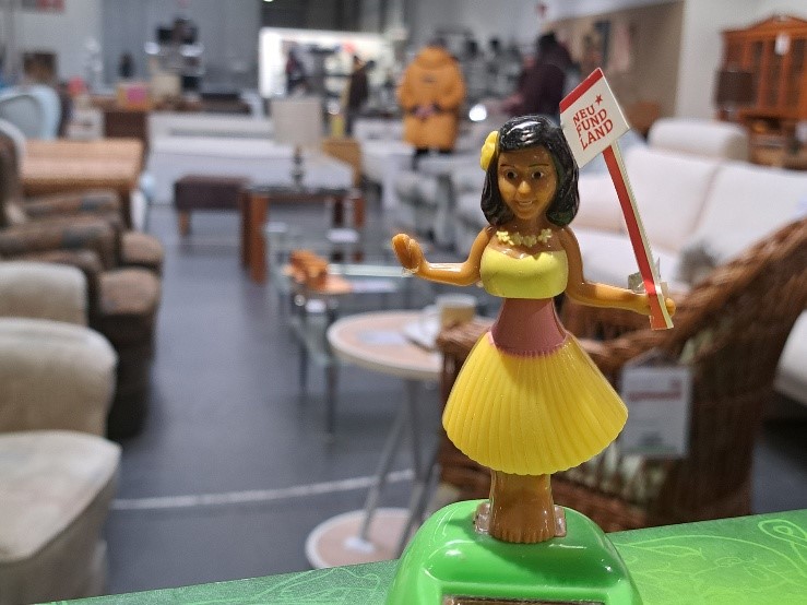 Plastikfigur mit schwarzen Haaren und einem gelben Sommerkleid die in der Hand ein Fähnlichen mit dem Logo des Secondhandwarenhaus Neufundland hoch hält. Im Hintergrund die Möbelabteilung des Warenhauses.
